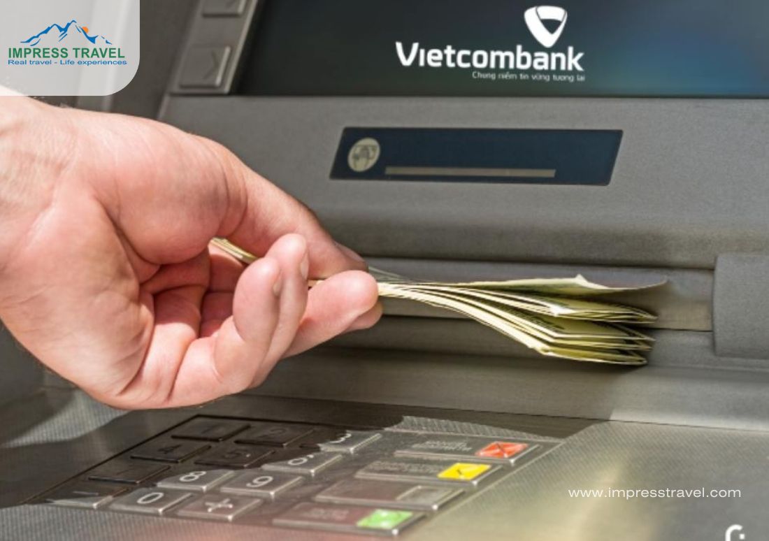 This image shows a close-up of a hand withdrawing banknotes from a Vietcombank ATM, focusing on the money and the keypad to highlight the transaction process. The ATM screen features the Vietcombank logo, linking the service to the bank. This common scene captures the routine of accessing cash, crucial for many transactions including currency exchange. The photograph, likely for promotional use by Impress Travel as indicated by the logo, emphasizes convenient banking solutions for travelers.