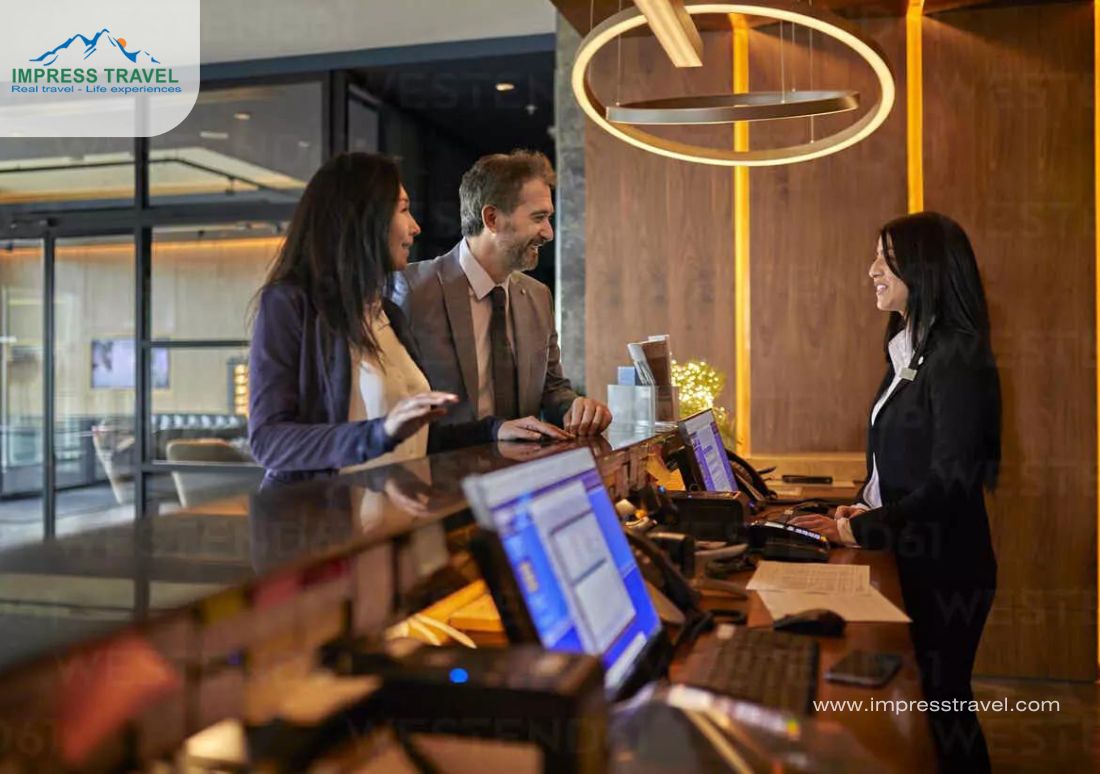 This image captures a cheerful interaction at a hotel reception, featuring a modern and warmly lit lobby with stylish decor. A receptionist in a black suit assists a couple, both dressed in business attire, who seem to be discussing hotel services or possibly exchanging currency—a common need for travelers. The presence of computers and various brochures on the counter suggests a well-equipped, customer-friendly environment that enhances the travel experience by offering essential services like currency exchange.